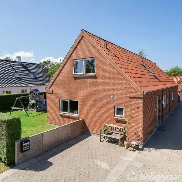 Rødt murstenshus omgivet af flisebelagt terrasse og træpergola. Møbleret med loungesæt og spisebordssæt med stole. Solrig omgivelse med klar blå himmel og grøn have i baggrunden.