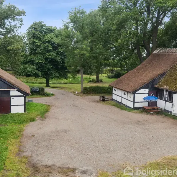 To længehuse med stråtag omringer en gårdsplads dækket af grus. Træer og græsplæner omkranser området, der skaber en landlig atmosfære.