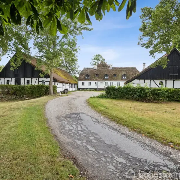 En traditionel stråtækt gårdbygning står omkranset af grønne træer og velplejede græsarealer. En snoet grusvej fører op til bygningen i et landligt område.