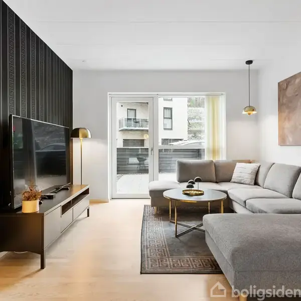 En grå sofa står i en moderne stue. Et sofabord er placeret foran, mens en stor fladskærm står på et TV-bord til venstre. Store vinduer bagved giver naturligt lys.