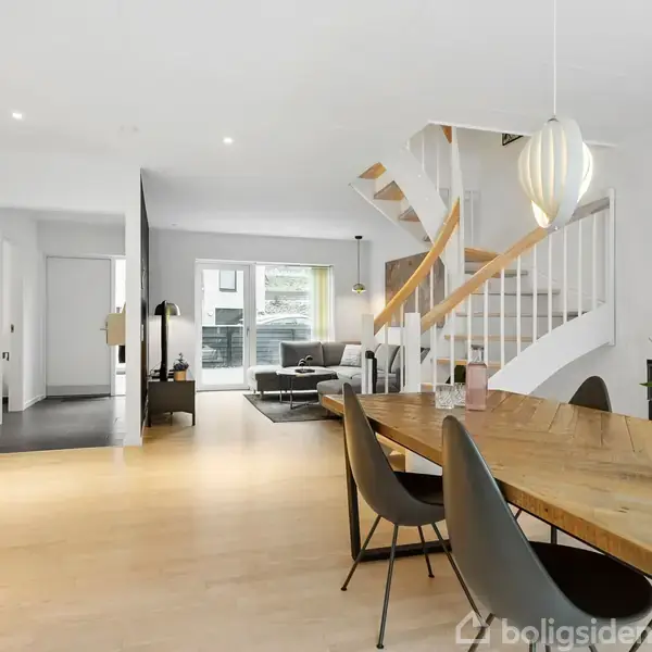 Stort spisebord med blomsterdekorationer står i et lyst stueområde. En trætrappe svinger opad mod højre. Sofaer og store vinduer findes længere inde i rummet. Ingen tekst til stede.