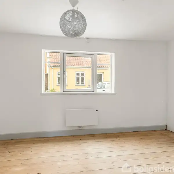 Et tomt værelse med trægulv og hvidmalede vægge, hvor en moderne lampe hænger fra loftet. Et vindue i baggrunden viser udsigt til en gul bygning udenfor.
