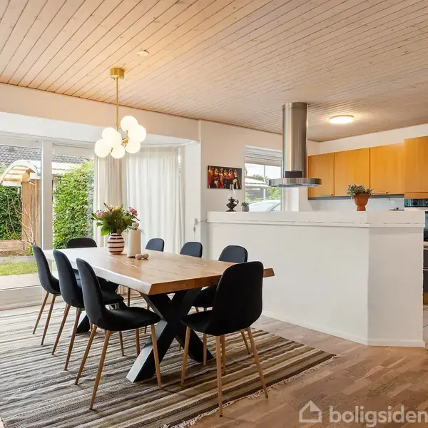 Spisebord med sorte stole står på et tæppe, omgivet af et lyst køkken med træloft. Store vinduer viser en grøn have udenfor, og en moderne lampe hænger over bordet.