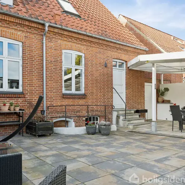 Rødt murstenshus med skråt tag omkranser flisebelagt terrasse. Havemøbler er arrangeret under en hvid baldakin til skygge. Trappe op til havedør. Ingen tekst synlig.