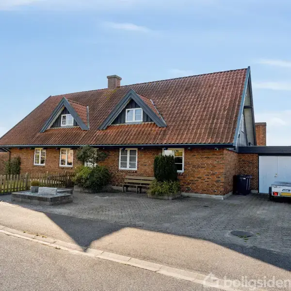 Murstenshus med rødt tegltag står på en flisebelagt indkørsel ved en vej. Omkring huset er der en lille have med buske og træer.