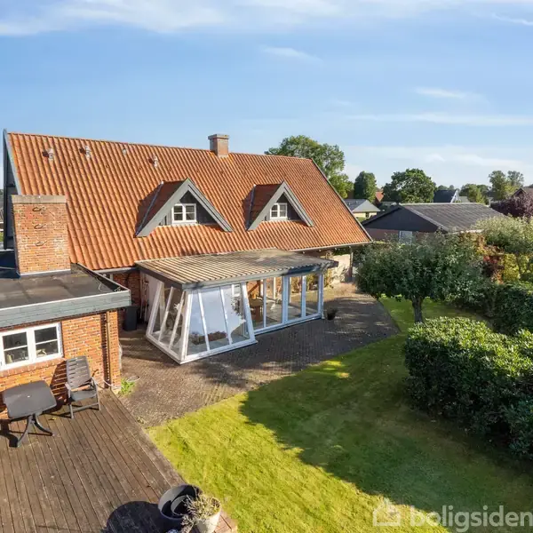 Et rødt murstenshus med skråt tag står i en grøn have med hække. En træterrasse med bord og stole er ved siden af en udestue med glaspartier.