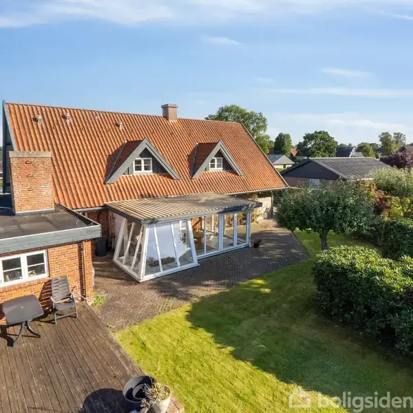 Et rødt murstenshus med en stejl taghældning har en udestue tilknyttet. I haven er der træterrasse med et cafésæt. Omkringliggende grønne buske og træer indrammer området.