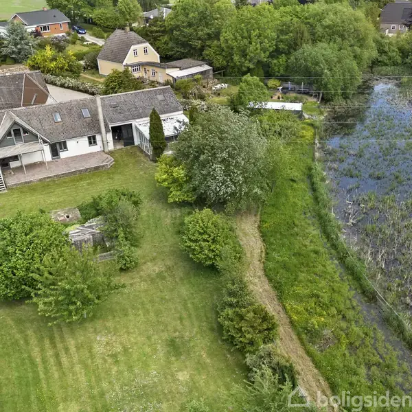 Et hvidt hus med tagterrasse ligger i en grøn have tæt på en sø. Omgivet af træer og buske i et villakvarter.