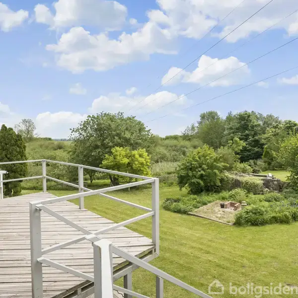 En træterrasse står stille med udsigt over en grøn have. Baggrunden indeholder buske, træer og en blå himmel med skyer.