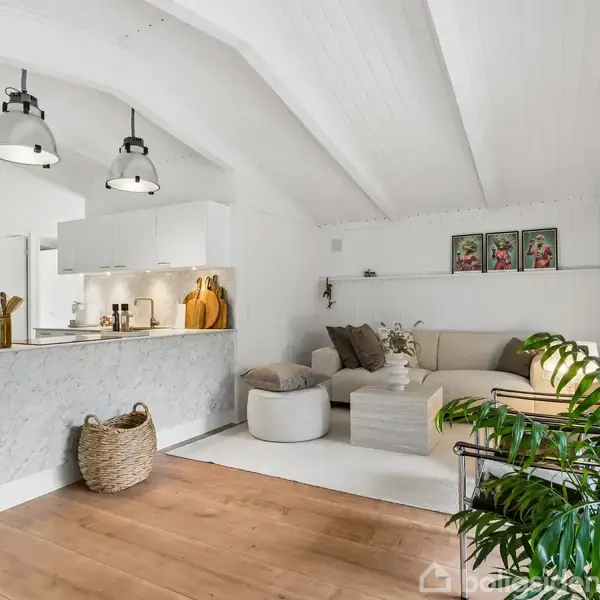 Et moderne køkken-alrum med en marmorø. To pendellamper hænger over øen. I baggrunden en beige sofa med puder. Grønne planter tilføjer liv. Hvidt loft med synlige bjælker.