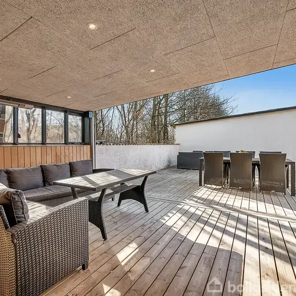 Rattan loungesæt står på en overdækket træterrasse, mens et spisebord med stole er placeret i sollys ved siden af. Høje træer og en hvid mur omgiver området.