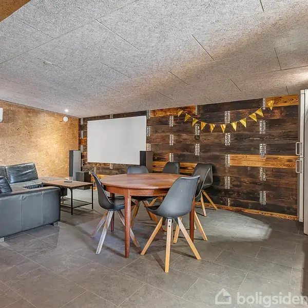 Sort lædersofa står i et moderne, minimalistisk rum med skråvægge. Et rundt træbord med sorte stole er i midten, og en bar med drikkevarer står ved siden af. Ingen tekst.