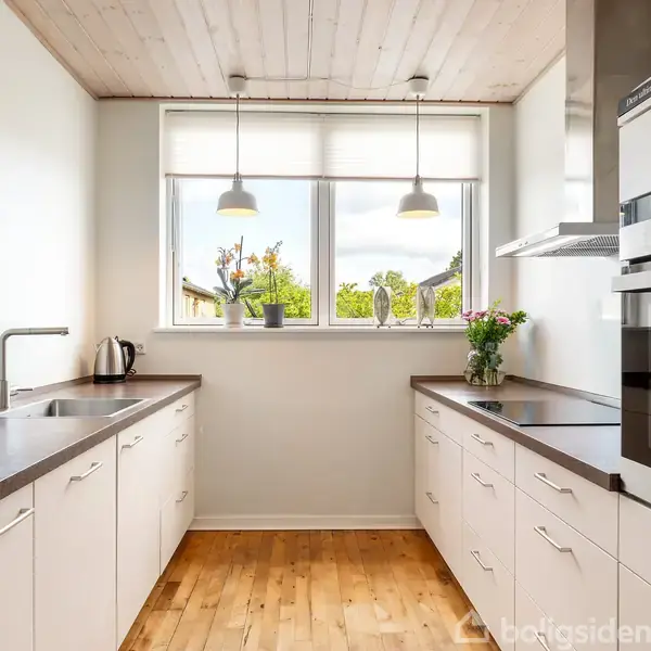 Et moderne køkken med hvide skabe, en sort bordplade, og integrerede hvidevarer. To hængelamper lyser rummet ved et vindue med udsigt til grønne træer. En kedel og blomsterpotte står fremme.