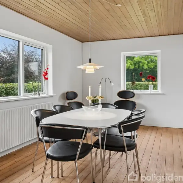 Et ovalt spisebord i træ med seks sorte stole står i et lyst rum. Hængelampe over bordet, store vinduer med udsigt til grøn have.