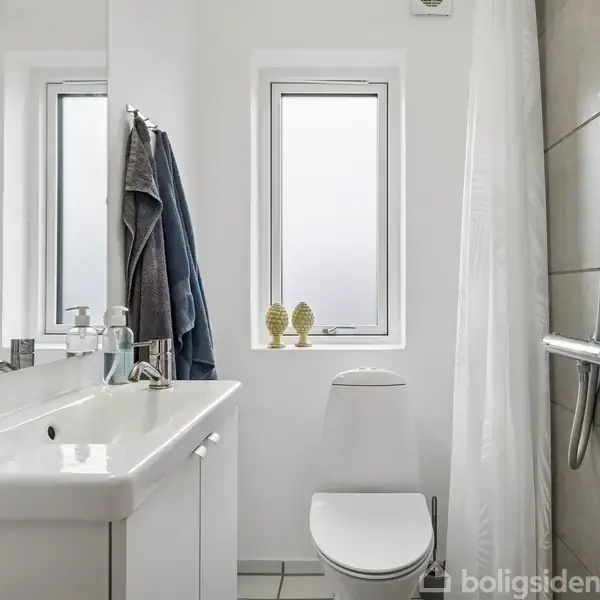 Hvidt badeværelse med vask og sæbedispenser til venstre. Bruser til højre. Toilettet står under et vindue med to dekorative figurer på karmen. Håndklæder hænger ved vasken.