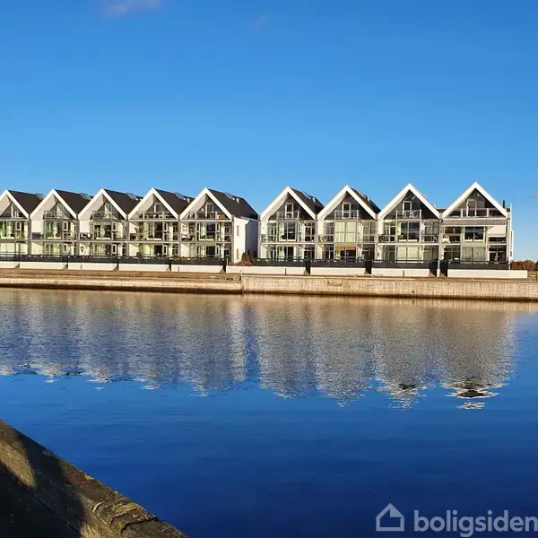 Rækkehuse med spidse tage står stille langs en kaj, reflekteret i det rolige vand under en klar blå himmel.