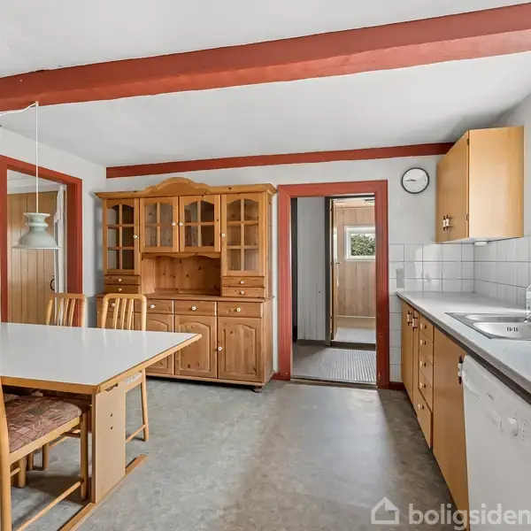 Et køkken med et spisebord og fire stole til venstre. Et stort træskab står ved væggen. Til højre er der en vask og opvaskemaskine under et vindue.