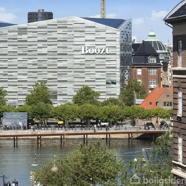 En stor, moderne bygning med teksten "Boozt" står ved en kanal. I forgrunden ses træer og en platform med mennesker, omgivet af ældre bygninger og åbent himmel.