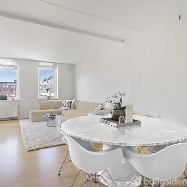 Et rundt marmorbord med hvide stole står i en lys stue. Ved siden af er en beige hjørnesofa foran vinduer med udsigt til bybygninger. En plante er placeret ved siden af sofaen.