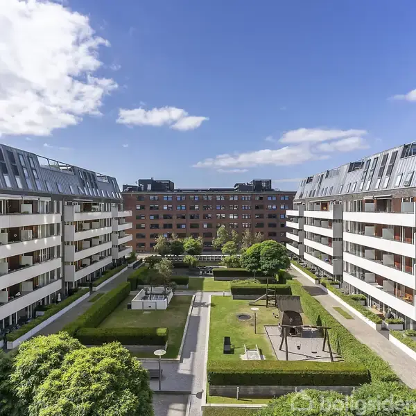 Boligblokke med altaner flankerer en grøn gårdhave med græsplæner, buske og en legeplads. Himlen er delvist dækket af skyer.