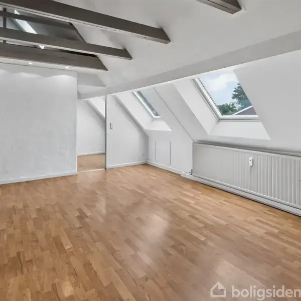 Loftet er et rum med skråvinduer, der slipper lys ind, og har en lys træparket. Vægge og loft er hvide med synlige bjælker. En radiator løber langs væggen.
