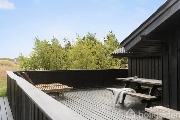 En træterrasse er udstyret med en bænk og et bord under et halvtag. Et tæppe er lagt på bænken, og en tom krus står på bordet. Skove omkranser området.