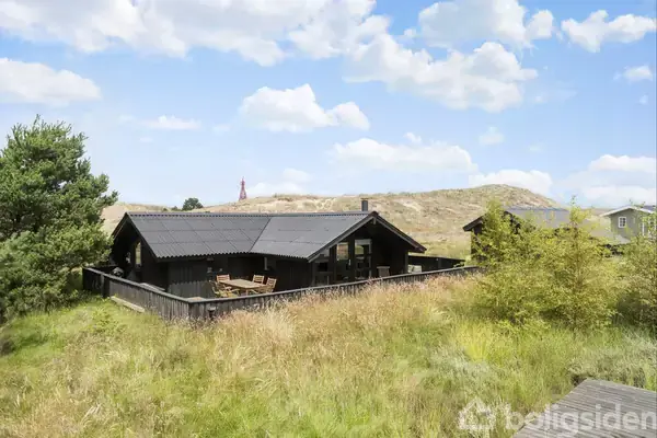 Et sort hus står i en grøn, bakket landskab. Foran huset er en terrasse med møbler. I baggrunden ses en rød-hvid antenne omringet af græsarealer og træer.