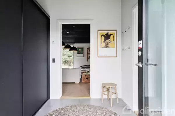 Døråbning fører til et værelse med hængende lamper, omgivet af lyse vægge og en skammel ved siden af en kunstplakat. Tæppe på gulvet, kroge på væggen.