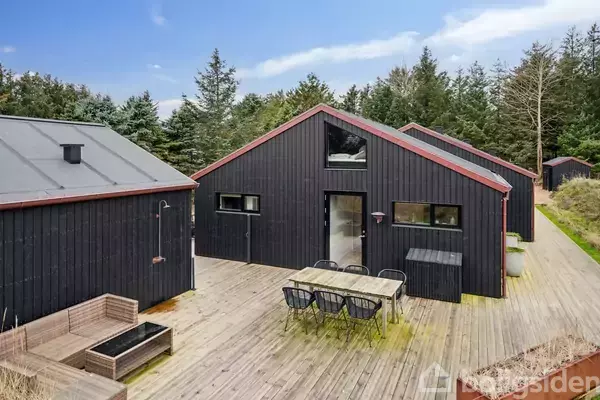 Et sort træhus står på en træterrasse, omgivet af skov. Der er et havebord med stole foran huset, og i baggrunden ses flere træbygninger.