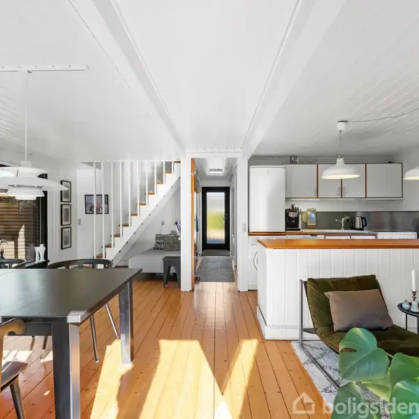 Spisebord står i et moderne køkkenalrum med trægulve. Pejs i murstensvæg til venstre. Trappe leder opad. Grønne lænestole ved siden af. Loftslamper hænger over spisepladsen.