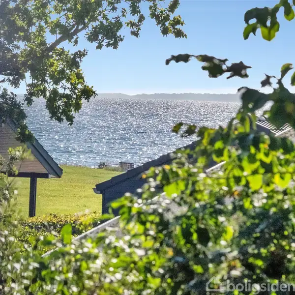 Et roligt hav skimrer i sollys, omgivet af grønne træer og tagene på husene i forgrunden, mens kysten kan ses i baggrunden.