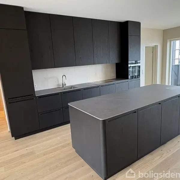 Sort køkken med moderne skabe og en køkkenø. Køkkenet er i en åben planløsning med lyst trægulv og store vinduer, der åbner ud til en balkon.