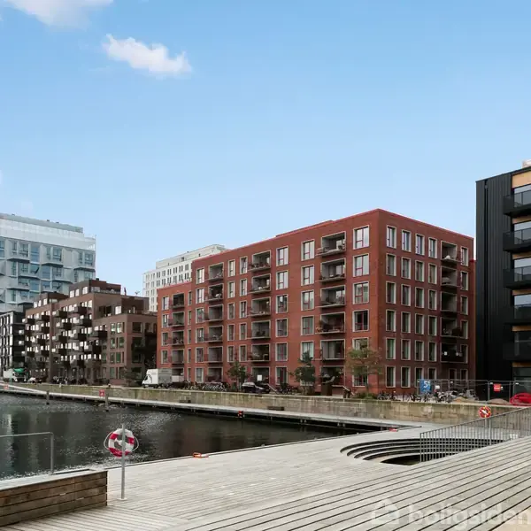 Flere moderne lejlighedsbygninger står langs en rolig kanal. Trædæk og redningskrans er placeret ved vandkanten. Himlen er overskyet med enkelte skyer.