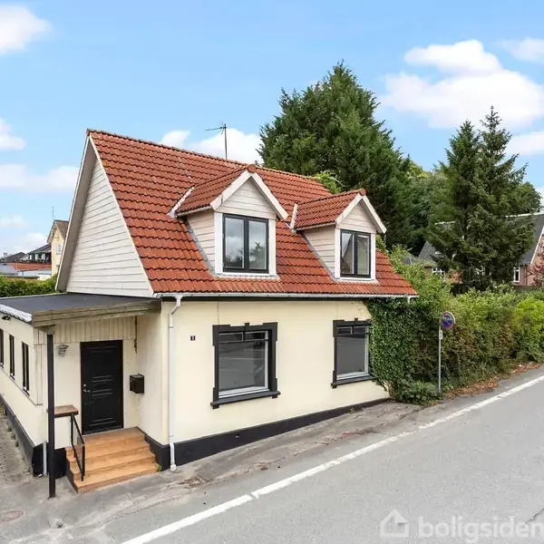 Hus med rødt tegltag og to kvistvinduer står ved vejkryds. Det har hvide vægge, sorte vinduer og en trædør med trapper. Hække og træer omkranser baggrunden på rolige villaveje.