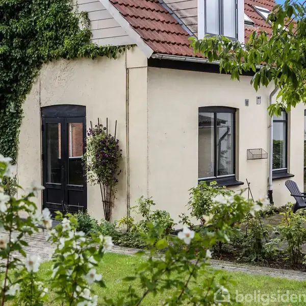 Et hvidt hus med sorte døre og vinduer står omgivet af grønne planter og blomster i en velholdt have. En stenterrasse med stole er synlig.