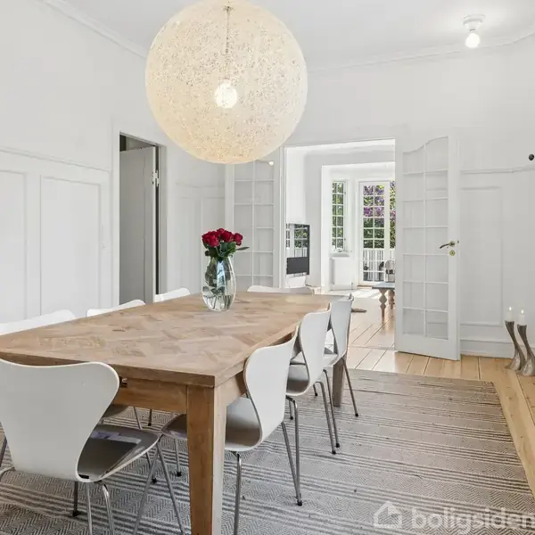 Et lyst spisestue med et træbord omgivet af hvide stole, en vase med røde roser på bordet. Store dobbeltglasdøre åbner til et tilstødende rum.