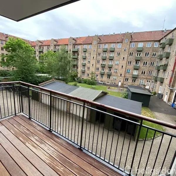 Balkon med træplanker står stille med udsigt over gårdområde og boligblokke. Omgivet af grønne træer og lejlighedsbygninger i baggrunden.