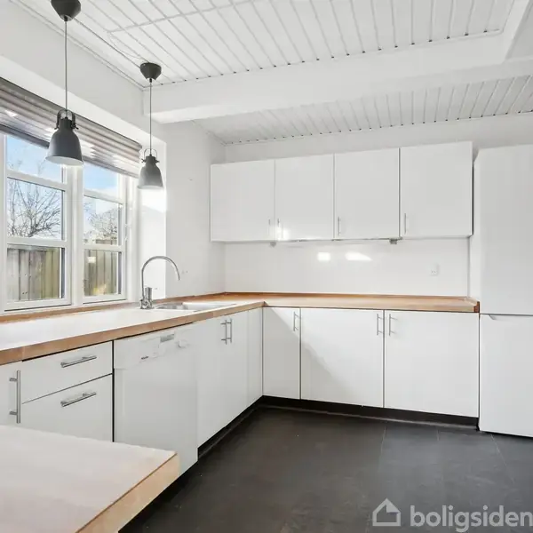 En moderne køkkenindretning med hvide skabe og træbordplader, udstyret med en vask. To hængende lamper oplyser rummet, der har et vindue med udsigt til en have.