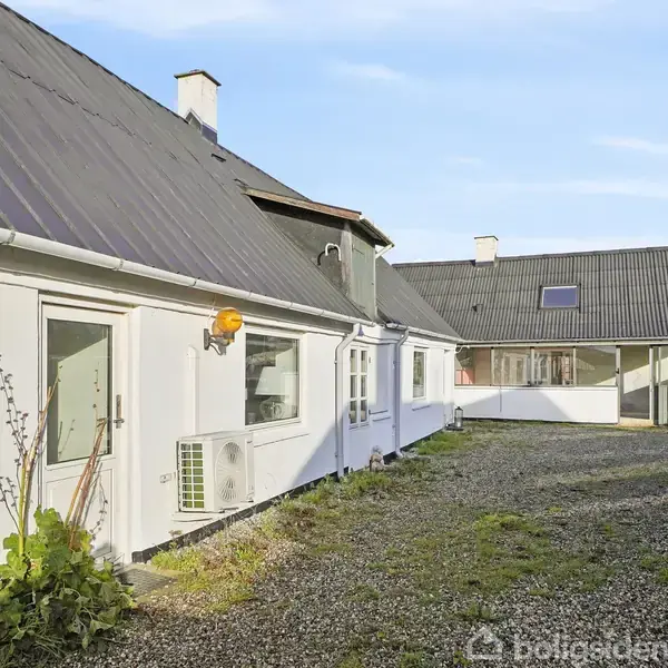 Hvidmalet hus med skråt tag, placeret langs grussti, omgivet af grønne planter. En gadelampe sidder tæt på døren. I baggrunden ses flere små bygninger.
