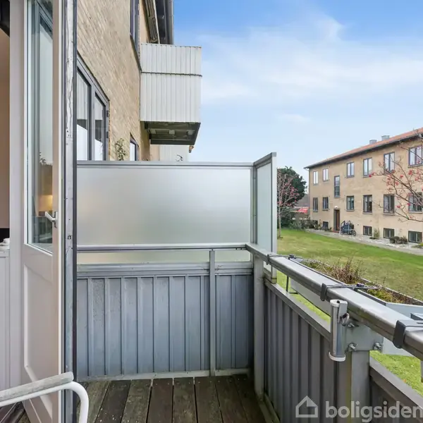 En lille altan har en åben dør til en stue med kunst på væggen; omgivet af beboelsesbygninger og græsplæner under en klar himmel.