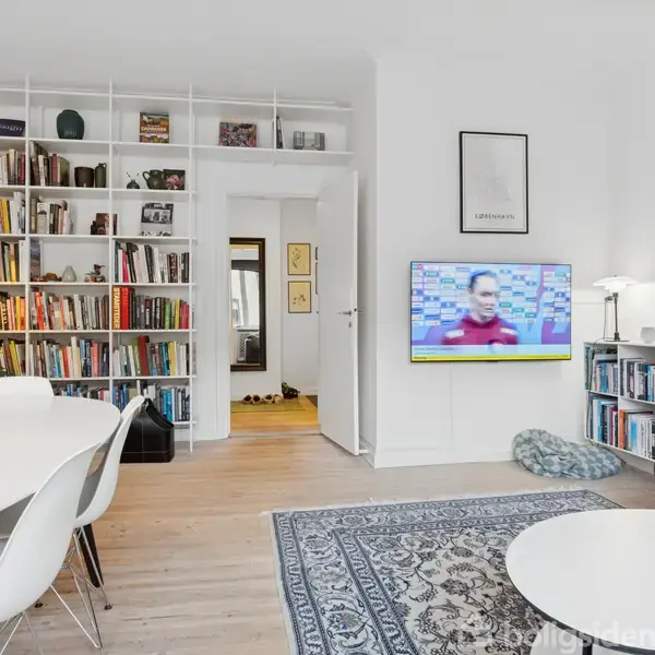 Et moderne stueområde indeholder et rundt spisebord med hvide stole. En vægmonteret TV viser en person i et interview. Indbyggede reoler fyldt med bøger dominerer bagvæggen.