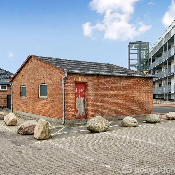 En enkeltstående rød murstensbygning med en falmet trædør omgivet af store sten på en asfalteret parkeringsplads. Til højre ses en moderne lejlighedsbygning i baggrunden.