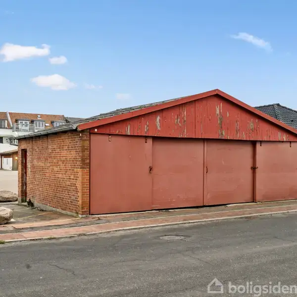 En ældre, rød garagebygning står tomt ved en vej. Bygningen er omgivet af parkeringsplads med store sten. I baggrunden ses en bygning med et skilt, hvorpå der står "Georgine".