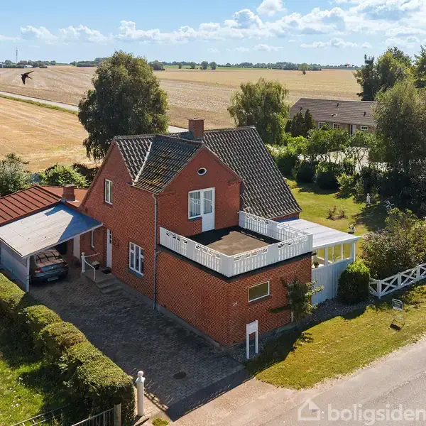 Rødt murstenshus med carport ved siden af, omgivet af have og landlig udsigt. Etage med terrasse ovenpå. Klart solskin oplyser scene med marker og træer i baggrunden.