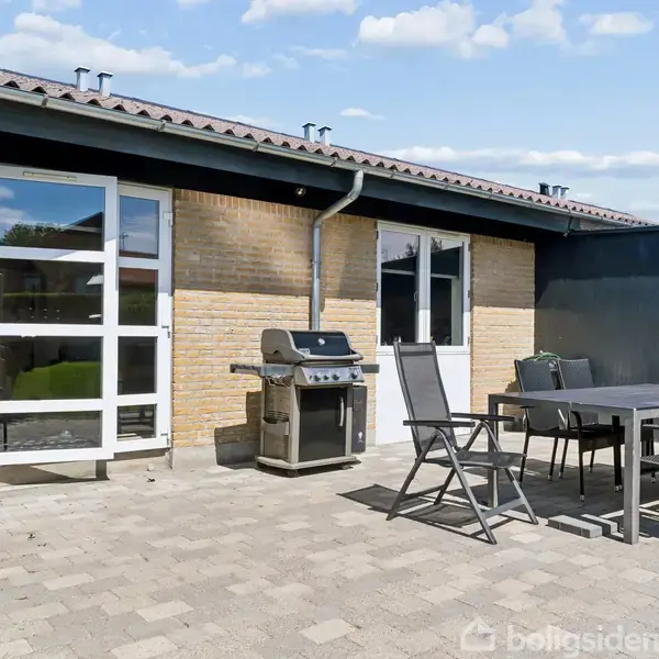 Terrasse med bord og stole placeret på fliser. En grill står ved husets murstensvæg under et skråt tag. Der er to vinduer og en dør til huset. Ingen synlig tekst.