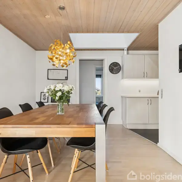 Blomster står på langbord i lys spisestue med seks sorte stole. En gylden loftslampe hænger over bordet. I baggrunden ses hvide skabe og en vægmonteret ur.