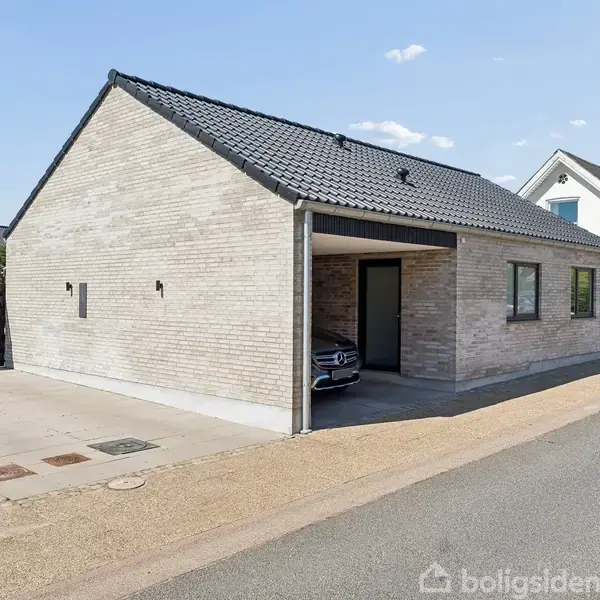 En moderne murstensvilla med sort tag er beliggende i et boligkvarter. En bil er delvis parkeret under et halvtag ved siden af huset. Klar himmel i baggrunden.