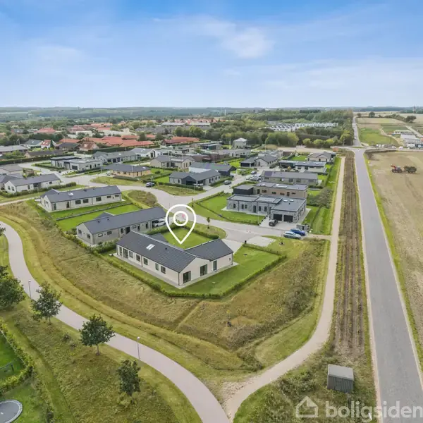 Et boligkvarter med moderne huse ses fra oven. En hvid markør fremhæver et bestemt hus. Græsarealer og veje omgiver kvarteret, der ligger i et åbent landskab.