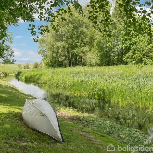En vendt kano ligger stationært ved et græsklædt område nær en stille flod omgivet af tætte grønne træer og buske under en blå himmel.