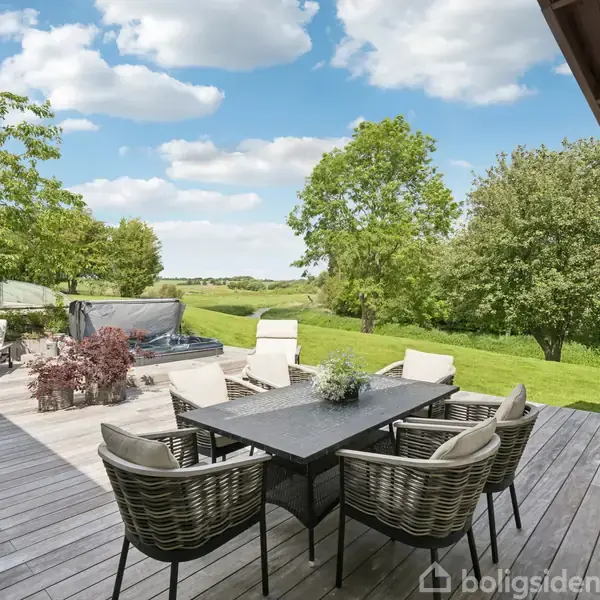 Terrasse med spisebord og seks stole, omgivet af grøn plæne og træer under en blå himmel med skyer. Luksusspa og liggestole i baggrunden.